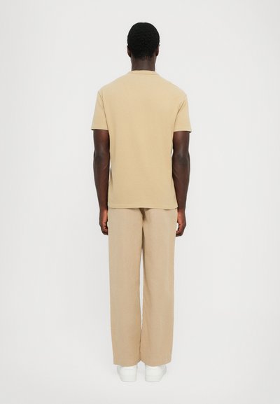 Beige kortärmad t-shirt matchad med beige vida byxor, båda i lättviktsmaterial. Modell vänd bort, bärande vita sneakers.