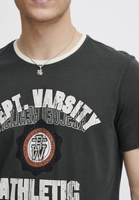 Tamno siva majica s okruglim izrezom, sa podebljanim belim tekstom i centralnim grafičkim emblemom. Srebrna lančić s privjeskom.
