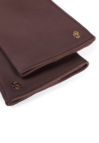 Un paio di guanti in pelle marrone scuro piegati con piccole decorazioni a forma di teschio dorato vicino ai polsi su sfondo bianco.