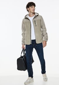 Beige bunda na zip s texturovanými fleecovými předními panely, šedými rukávy a kapucí. Nosit s tmavě modrými džíny a světle šedými teniskami. Černá taška v ruce.