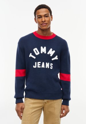 Młody mężczyzna w granatowym swetrze z czerwonym kołnierzem i rękawami, "Tommy Jeans" tekst, oraz beżowych spodniach, stojący na białym tle.