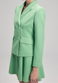 Mátově zelený blejzr s dvouřadým designem, texturovanou tkaninou, klopami a dvěma předními kapsami, v kombinaci s odpovídajícími rozšířenými sukněmi.