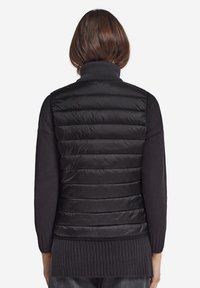 Schwarze Pufferweste mit hohen Kragen, die horizontale Stepppanelen, eine glatte Textur und ein tailliertes Design über einem dunklen Pullover aufweist.