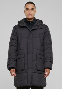 Crna podstavljena parkasta jakna s kapuljačom, koja ima dva prednja džepa i zatvaranje na zip. Materijal izgleda glatko s šivenim dijelovima za izolaciju.