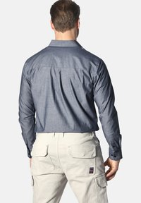 Herren Langarmhemd aus dunkelgrauem Chambray-Stoff, mit klassischem Kragen und Schulterfalten. Kombiniert mit hellen Cargo-Hosen.