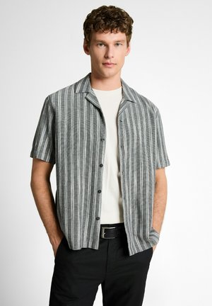 Junger Mann mit lockigem Haar, der ein grau-weiß gestreiftes kurzärmeliges Hemd über einem weißen T-Shirt und einer schwarzen Hose trägt.