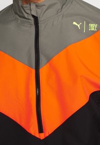 Grå, orange och svart jacka med dragkedja framtill, med Puma och First Mile-logotyper på övre högra bröstet.