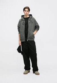 Hanorac gri cu fermoar, cu două buzunare frontale, purtat cu un tricou negru și pantaloni cargo negri. Întreaga ținută este completată cu cizme verde-maro.