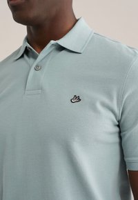 Lichtblauw poloshirt van gestructureerde stof met een klassieke kraag, een knoopsluiting met twee knopen en een klein zwart geborduurd vogellogo op de borst.