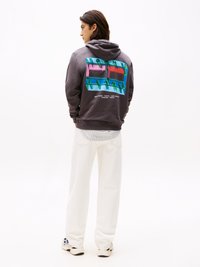 Hanorac gri cu un design grafic colorat pe spate, având textul "TOMMY JEANS" și culori vibrante. Asortat cu pantaloni albi.