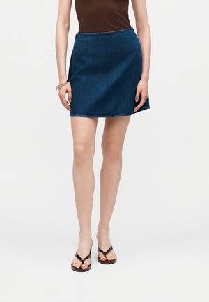 Žena nosící tmavě modrou džínovou mini sukni, hnědý top bez rukávů, černé sandály s tenkými pásky a zlatý náramek na levém zápěstí.