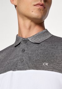 Grå og hvit polo-skjorte med krage, tre knappelukkinger og en liten brodert CK-logo på brystet. Myk, strukturert stoff.