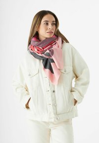 Femme portant une veste en denim crème et un pantalon avec une écharpe à motifs multicolores rose, rouge et gris, regardant sur le côté avec les mains dans les poches.