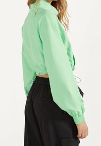 Kort jacka i klar mintgrön, med krage, elastiska ärmmuddar och justerbar dragsko i midjan. Tillverkad med svarta byxor.