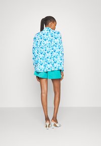 Blusa com padrão floral em azul claro com flores em azul mais escuro, cintura ajustada e mangas longas. Combinada com calções em teal e sabrinas brancas de salto.