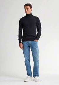 Marinblå polotröja med figurnära design och långa ärmar, kombinerad med ljusblå jeans och vita sneakers mot en neutral bakgrund.