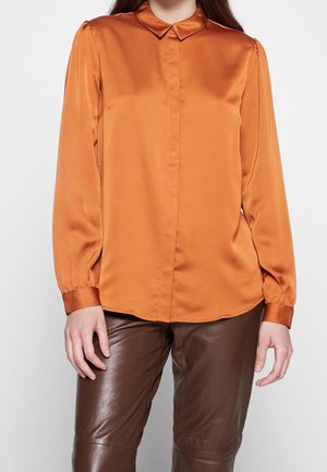 Kvinde iført en brændt orange langærmet bluse med krave og brune læderbukser, stående mod en ensfarvet baggrund.