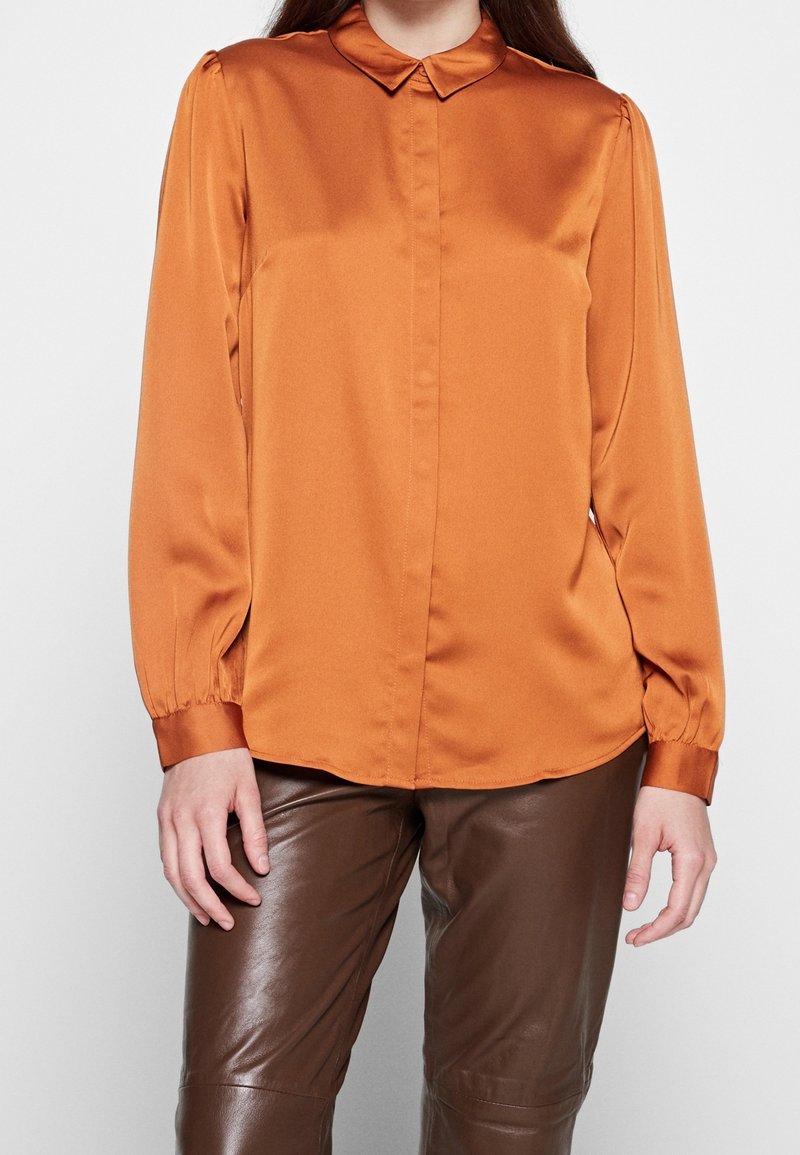 Femme portant un chemisier à manches longues orange brûlé avec un col et un pantalon en cuir marron, debout devant un fond uni.