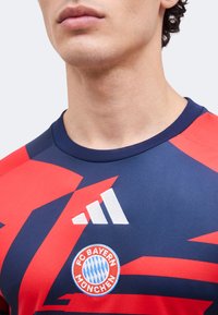 Lähikuva henkilöstä, joka on pukeutunut tummansiniseen ja punaiseen FC Bayern Münchenin jalkapallopaitaan, jossa on Adidas-logo rinnassa.
