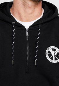 Felpa nera con zip, dotata di una toppa con logo bianco e laccetti testurizzati con un motivo in bianco e nero. Tessuto morbido, vestibilità rilassata.