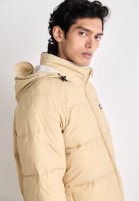 Beige puffer jacket with quilted design, adjustable hood, and front zipper. Mixed fabric texture with a label on the chest.