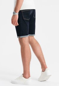 Bermudas de mezclilla en azul oscuro con dobladillos rematados. Presenta bolsillos traseros y costuras amarillas visibles. Combinadas con zapatillas blancas.
