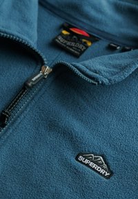 Chaqueta de forro polar azul con cremallera negra parcialmente cerrada y logo bordado de Superdry con diseño de montaña en el pecho.