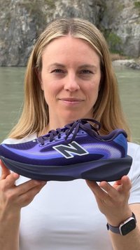 Γυναίκα με λευκό πουκάμισο κρατάει ένα μωβ και μαύρο αθλητικό παπούτσι New Balance με φόντο βραχώδη νερά.