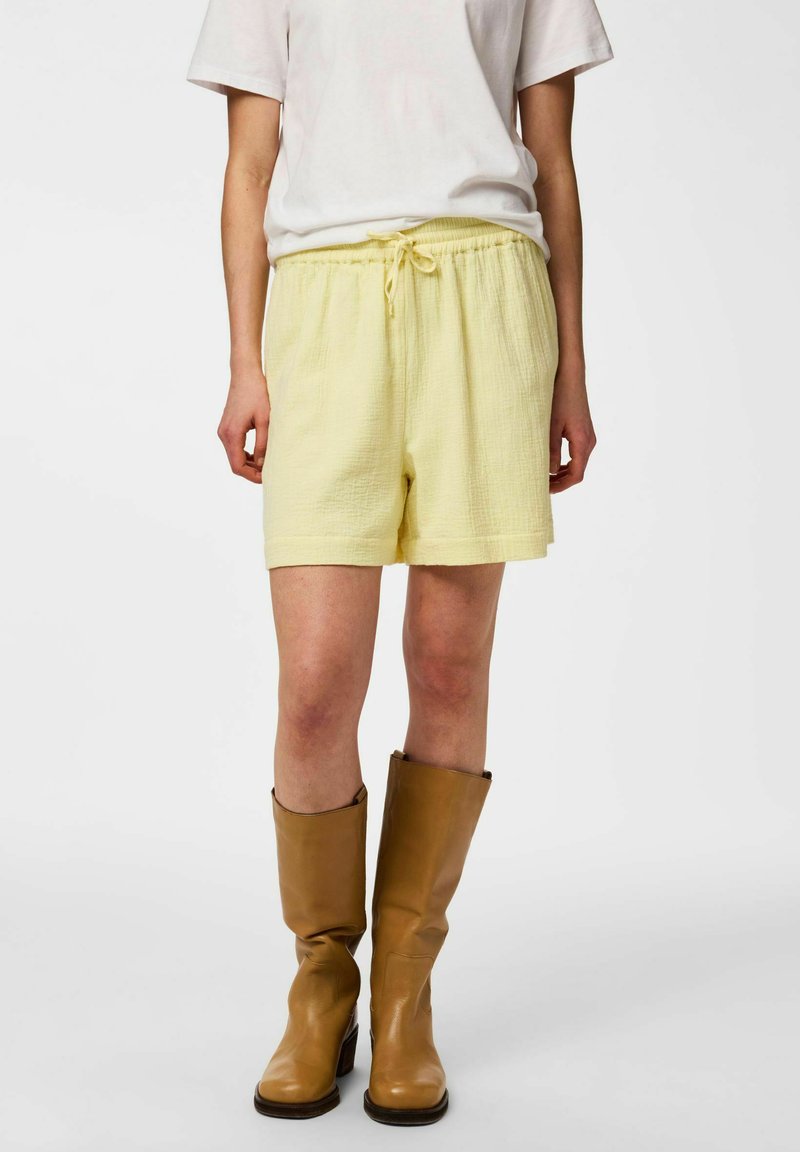 Personne portant un short texturé jaune clair avec taille à cordon, une chemise blanche à manches courtes et des bottes en cuir marron mi-mollet.