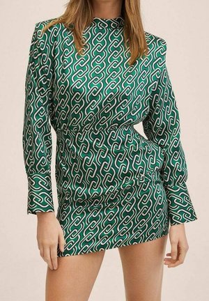 Vrouw draagt een groene, lange mouwen, korte jurk met een wit-zwart schakelpatter en staat met de handen bij de heupen.