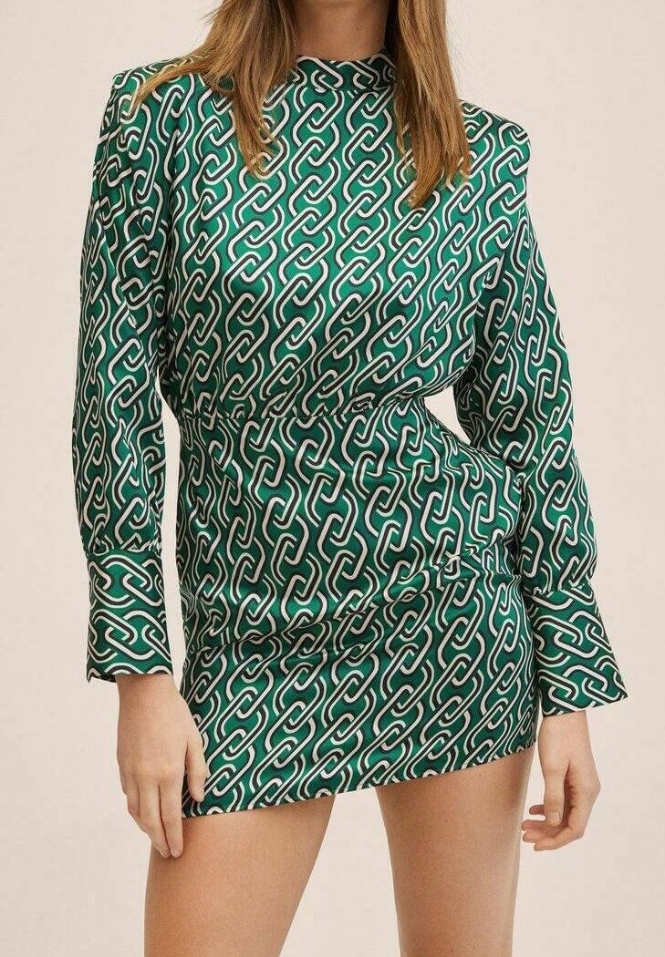 Femme portant une robe courte verte à manches longues, avec un motif de chaînes blanches et noires reliées, debout les mains près des hanches.