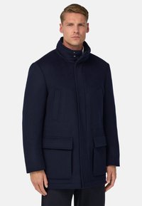 Homme portant un manteau à col montant navy foncé avec des poches à rabat à l'avant et des poches poitrine à fente verticales, se tenant devant un fond blanc uni.