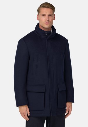 Muž oblečený v tmavě modrém vysokém límci s kapsami na klopě a vertikálními kapsami na hrudi, stojící před jednoduchým bílým pozadím.