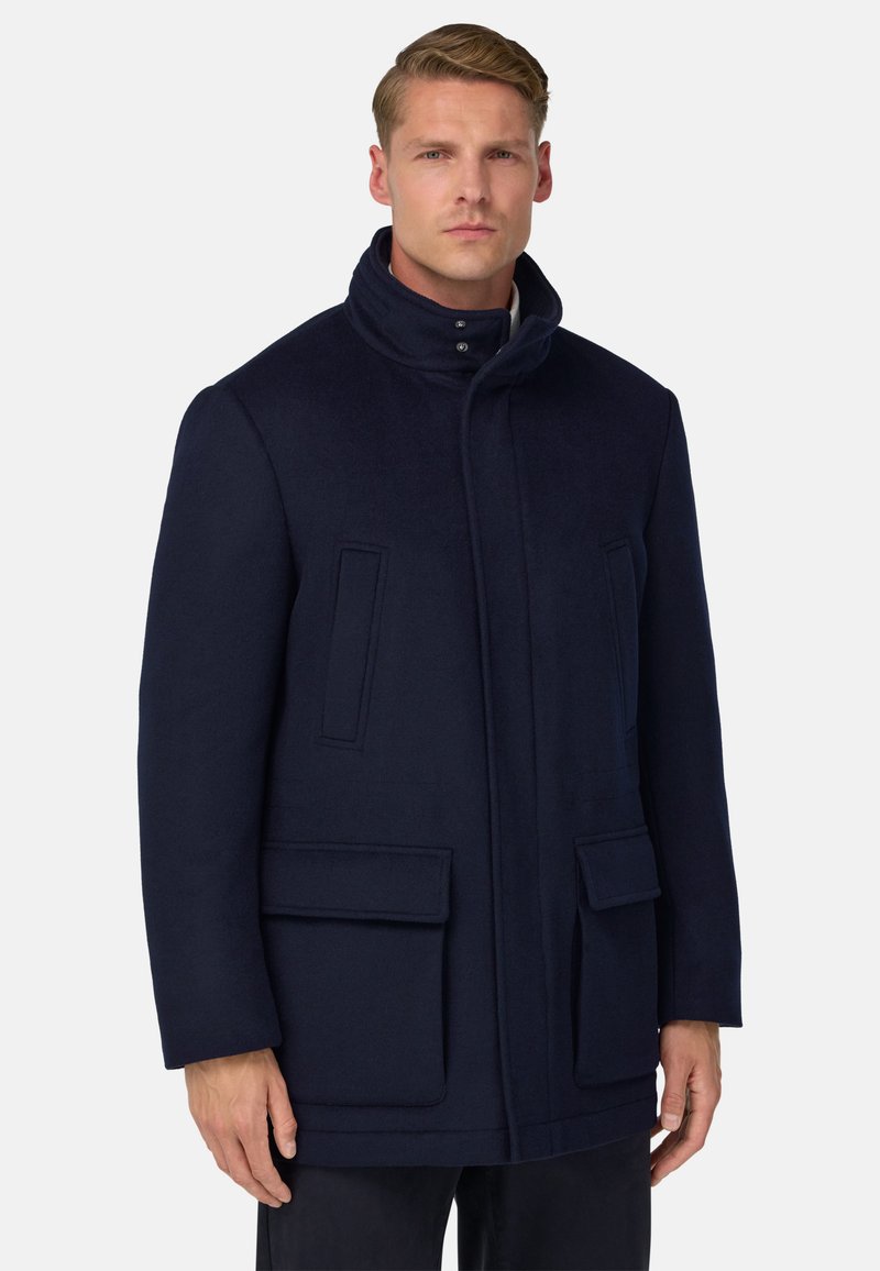 Homme portant un manteau à col montant navy foncé avec des poches à rabat à l'avant et des poches poitrine à fente verticales, se tenant devant un fond blanc uni.