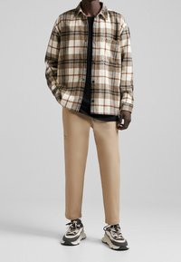Koszula flanelowa w kratę w odcieniach brązu, kremu i beżu; czarna koszulka pod spodem; beżowe zwężane spodnie; czarno-białe trampki z grubymi podeszwami.