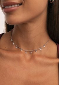 Collana in argento con piccoli ciondoli ovali pendenti indossata al collo e sul clavicollo di una donna, su uno sfondo neutro.