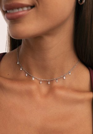 Collarín de cadena de plata con pequeños charm ovalados colgantes, lucido en el cuello y la clavícula de una mujer sobre un fondo neutro.