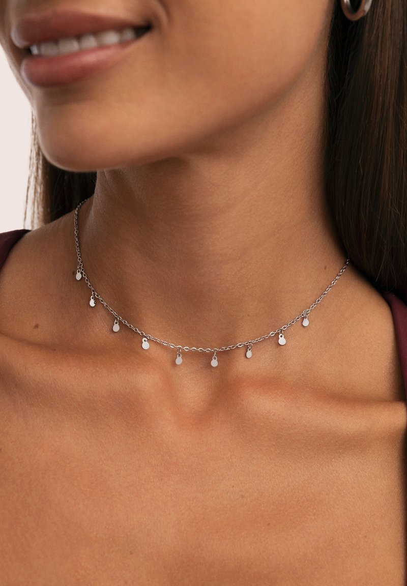 Collana in argento con piccoli ciondoli ovali pendenti indossata al collo e sul clavicollo di una donna, su uno sfondo neutro.