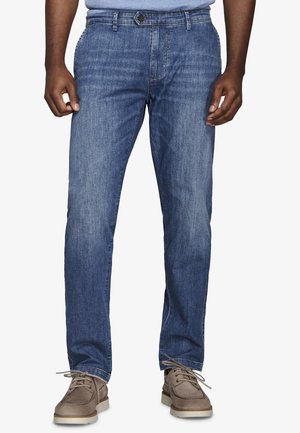 Mann trägt mittelblaue Jeans mit geradem Bein und hellbraune Schnürschuhe, steht mit entspannt an den Seiten hängenden Händen.