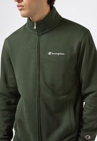 Man in een donkergroen Champion jack met rits, met een wit logo op de borst en een klein logo op de linkermouw.