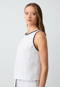 Haut sans manches en lin blanc avec bord noir, présentant une coupe décontractée et une texture subtile. Conçu avec un col haut et un ourlet droit.