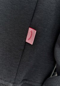 Temná sivá tkanina s hladkým povrchom, na ktorej je pripevnená malá ružová visačka s dizajnom rias.