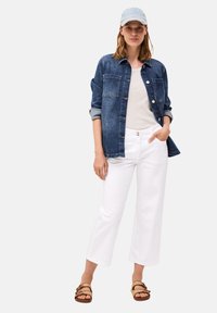 Camisa de denim en azul oscuro con detalles de botones, superpuesta sobre una blusa de color claro. Pantalones blancos de pierna ancha y sandalias trenzadas en color tostado completan el conjunto.