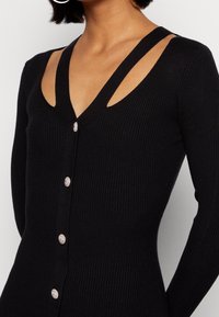 Maglione nero a coste con scollo a V profondo, caratterizzato da spalline con tagli e cinque bottoni decorativi sul davanti. Tessuto morbido in tutta la sua superficie.