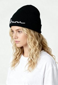 Zwarte gebreide muts met geribbelde textuur en wit cursief "Amour" geborduurd aan de voorkant, gecombineerd met een effen wit t-shirt.