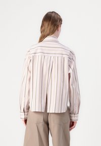 Stripet skjorte i lys rosa med hvite og gule striper. Har plisserte detaljer bak og oppbrettede ermer. Tøyet ser glatt ut.
