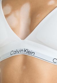 Nærbilde av en person som har på seg en hvit Calvin Klein bralette med logo på underbåndet.