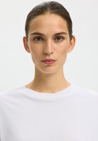 Woman with slicked-back hair and light makeup wearing a plain white crew neck shirt against a plain light background.