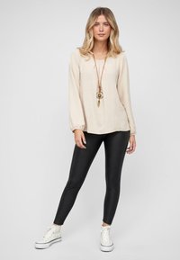 Beige textuur long-sleeve top met een ronde hals, gecombineerd met zwarte leggings en witte sneakers. Accessoire omvat een gelaagde ketting.