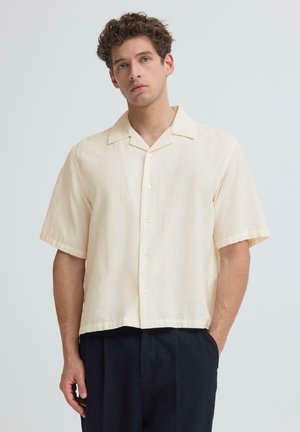 Junger Mann mit lockigem Haar, der ein cremefarbenes Kurzarmhemd mit Knöpfen und dunkle Hosen vor einem einfarbigen hellen Hintergrund trägt.